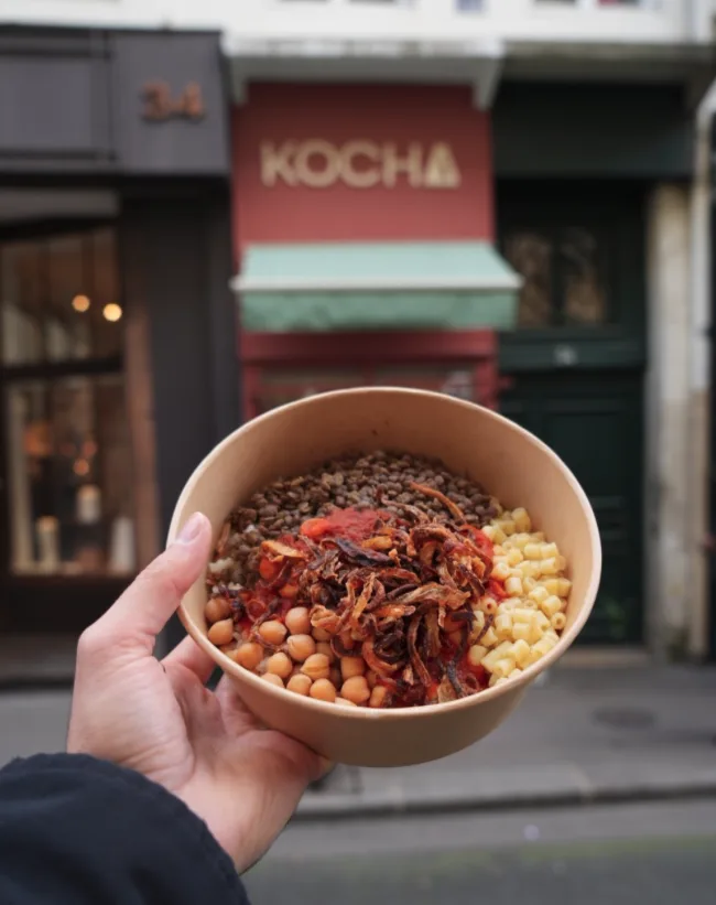 Un bol de kochari KOCHA — lentilles, pois chiches, pâtes, riz, oignons frits et sauce tomate — tenu devant la devanture du restaurant au 34 Rue Coquillière à Paris